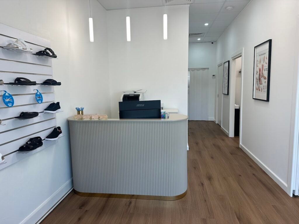 Front reception desk and waiting area of a modern podiatry clinic with wooden floors and a wall display of orthopedic shoes.