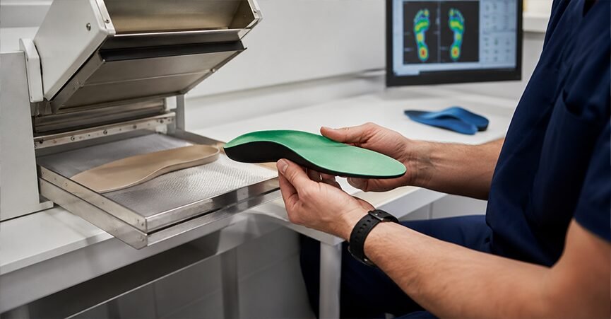 A podiatrist holding a green heat-moldable orthotic shell next to a molding machine, with a foot pressure map displayed on a monitor in the background.